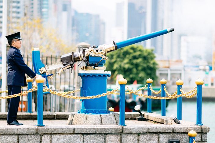 A guard in dark uniform and peaked cap standing at the breech of the polished Noonday Gun on its blue mounting, inside a low granite enclosure with blue posts and gold chains, the towers of Causeway Bay blurred behind