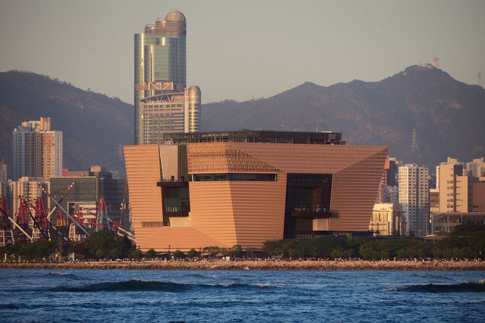 The bronze-clad Hong Kong Palace Museum, wider at the top than at its base, catching low sun on the West Kowloon waterfront, harbour water in the foreground and the tall ICC tower and green hills behind