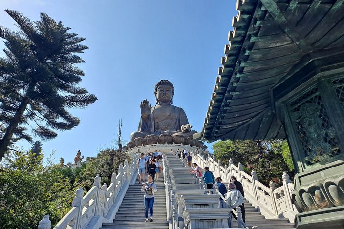 Visitors climbing a wide balustraded stone stairway towards the Tian Tan Buddha, the seated bronze figure with its right hand raised on a lotus at the top, a bronze pavilion roof at the right and pines at the left under a clear blue sky