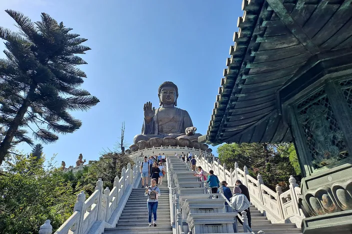 Visitors climbing a wide balustraded stone stairway towards the Tian Tan Buddha, the seated bronze figure with its right hand raised on a lotus at the top, a bronze pavilion roof at the right and pines at the left under a clear blue sky