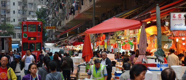 A crowded street market in North Point with a red double-decker tram edging along the tram tracks that run down the middle of the street, canvas awnings and hanging red lanterns on both sides, stacked crates and shoppers filling the roadway, tenement blocks rising above