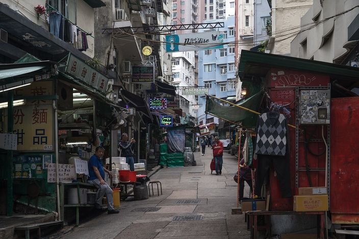 A narrow, sloping Hong Kong side lane off Elgin Street, with old noodle-shop signage, hanging laundry, a green neon 'Paradise' sign and a man seated among stacked crates, tenements rising behind.