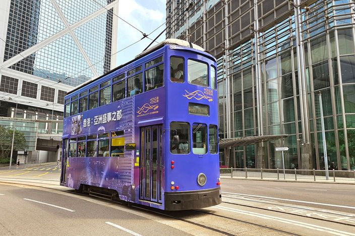 A purple double-decker "ding ding" tram runs along an empty road between glass-and-steel office towers in Central, Hong Kong Island, in bright daylight