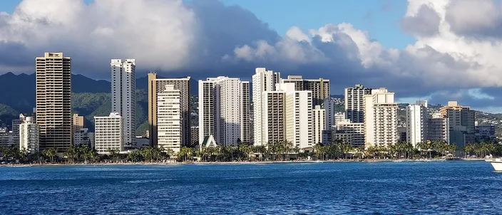 Waikīkī from the water: a wall of white hotel towers rising straight off a strip of pale sand under cumulus, the green ridges of Oʻahu behind them