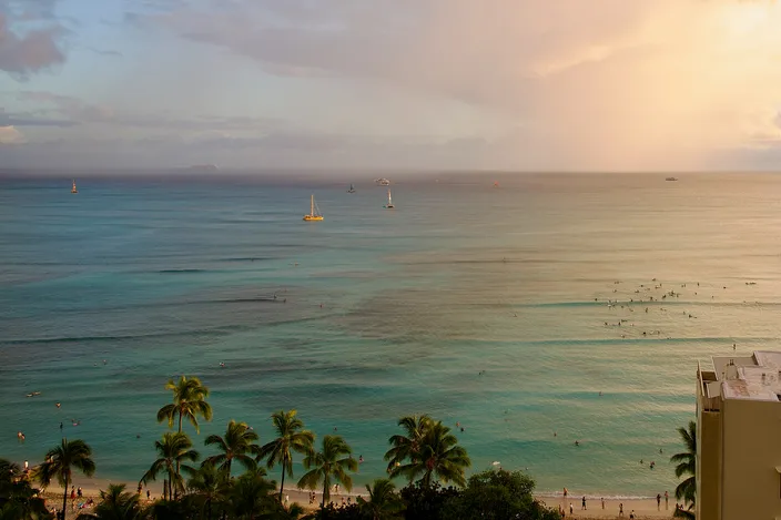 Waikīkī from above at the end of the day: the pale shelf of the reef under turquoise water, surfers waiting at the breaks far out, sailboats beyond, palms and a strip of sand along the bottom of the frame