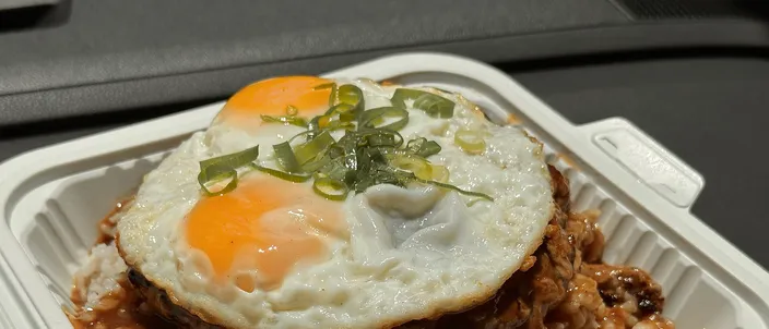 A loco moco in an open white takeout box: two sunny-side-up eggs topped with sliced green onion over a hamburger patty, on white rice soaked in brown gravy
