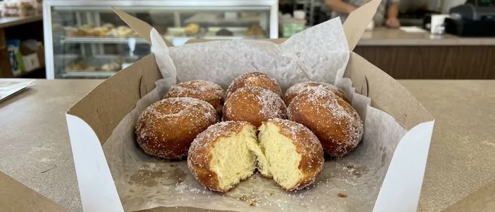 AI-generated image of plain malasadas rolled in granulated sugar in an open white bakery box, one torn open to show the soft interior