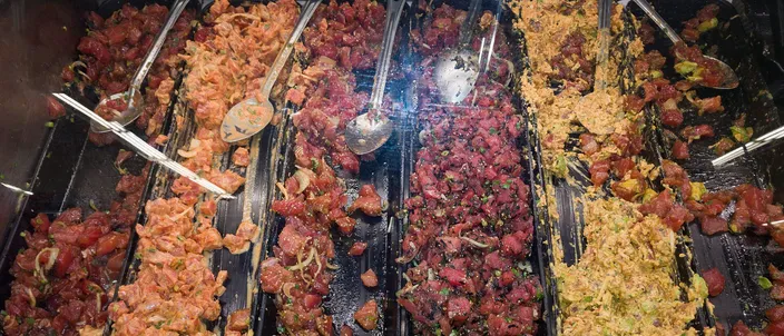 A supermarket poke case in Hawaiʻi: long trays of cubed raw tuna and salmon in shoyu, spicy mayonnaise and seaweed dressings, each with a serving spoon
