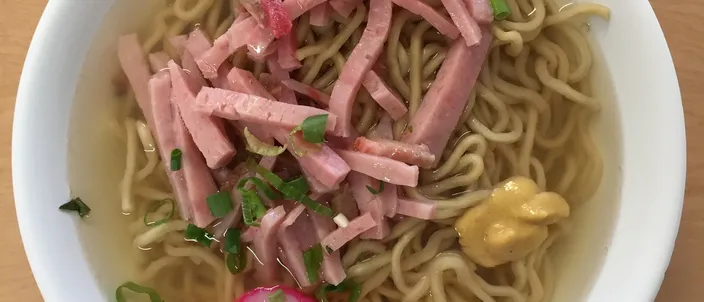A white bowl of saimin seen from above: thin yellow noodles in clear broth under strips of pink ham, sliced green onion, a dab of mustard and a swirled kamaboko fishcake round