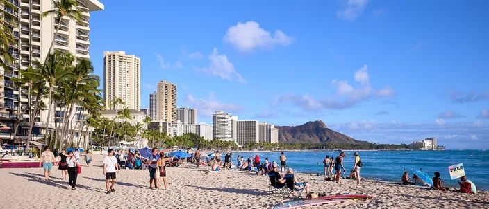 Waikīkī Beach on a clear day, sunbathers and walkers on wide pale sand, hotel towers and palms along the left and Diamond Head's ridge at the end of the shore