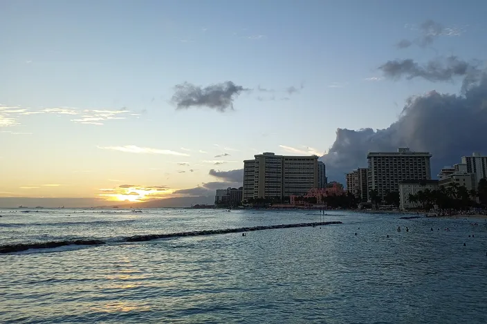 Sunset over Kūhiō Beach in Waikīkī, the low sea wall running out across calm water toward hotel towers on the shore