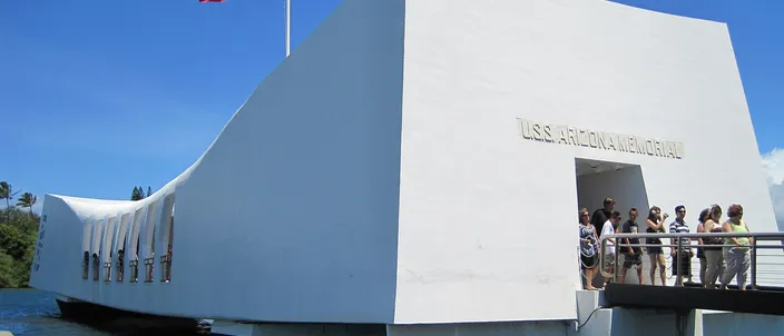The white USS Arizona Memorial over the water of Pearl Harbor, visitors at its open doorway and the US flag flying above it