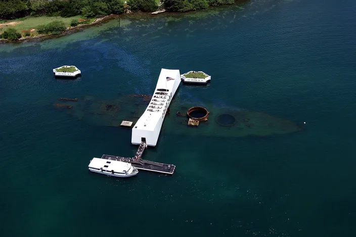 Aerial view of the white USS Arizona Memorial spanning the dark outline of the sunken battleship in the green water of Pearl Harbor, a launch moored beside it