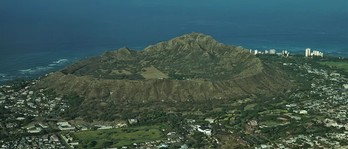 Diamond Head seen from the air: the broad green crater rim rising above the houses of eastern Honolulu, the ocean beyond it