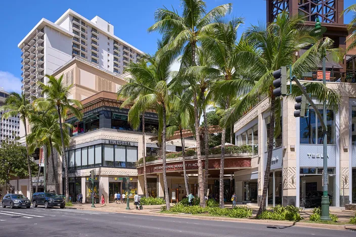Kalākaua Avenue in Waikīkī under tall palms, a hotel tower and low shopping arcades lining the street on a sunny morning