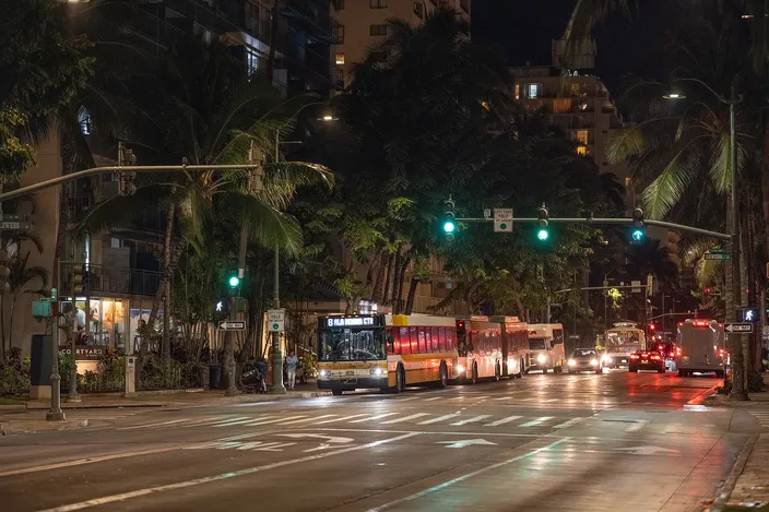 TheBus buses lined up along Kūhiō Avenue in Waikīkī at night under palms and street lights