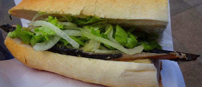 A balık ekmek: a fillet of grilled fish in a split length of white bread with raw onion and green leaves, held on white paper.