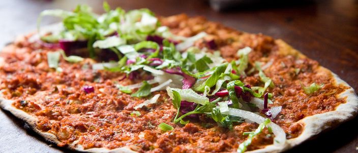 A single thin round lahmacun on a dark plate, its surface spread with a thin layer of minced meat and scattered with green leaves.