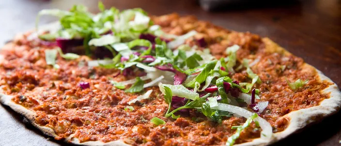 A single thin round lahmacun on a dark plate, its surface spread with a thin layer of minced meat and scattered with green leaves.
