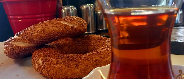Two sesame-crusted simit rings on a saucer beside a tulip-shaped glass of dark Turkish tea on a table.