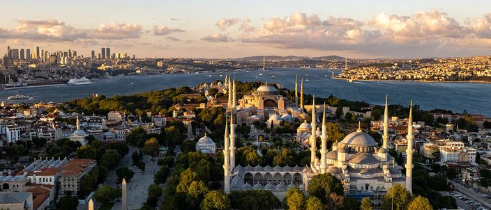 Aerial golden-hour view over the historic peninsula: the Blue Mosque and Hagia Sophia among trees with the Bosphorus, a suspension bridge and the modern skyline behind.