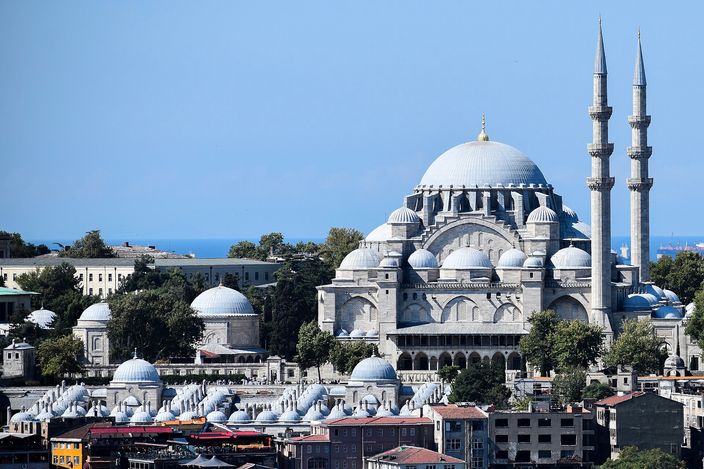 The Süleymaniye Mosque on its hilltop, a cascade of pale grey domes and half-domes with two minarets against a clear blue sky, smaller domed tombs and city rooftops below and open water on the horizon.