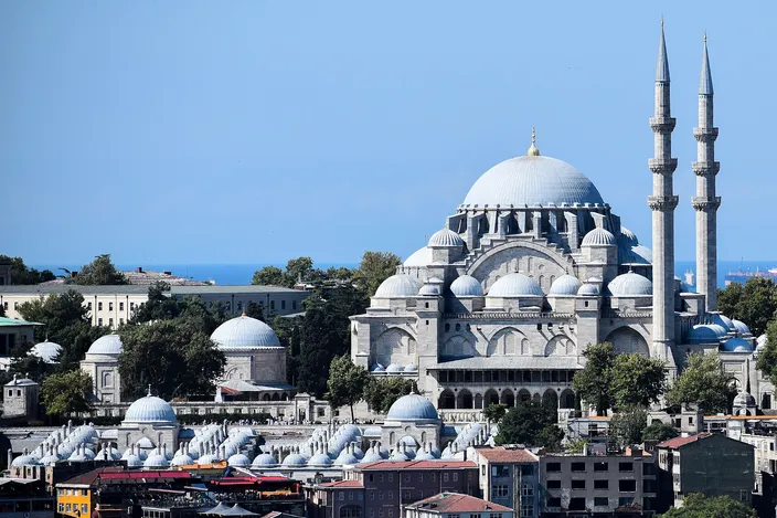 The Süleymaniye Mosque on its hilltop, a cascade of pale gray domes and half-domes with two minarets against a clear blue sky, smaller domed tombs and city rooftops below and open water on the horizon.