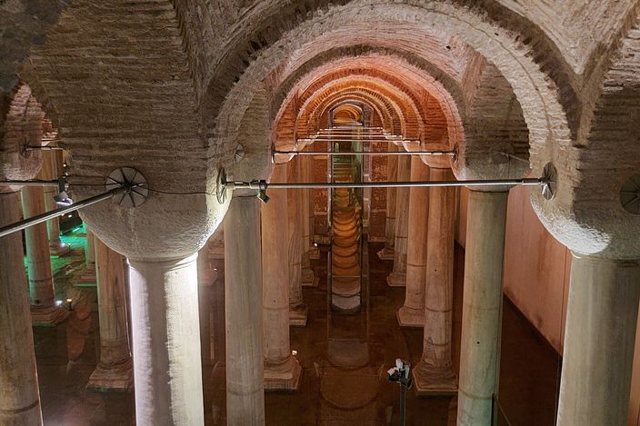 Ranks of columns standing in shallow water beneath brick barrel vaulting in a dimly lit underground cistern, warm light on the far vaults and long reflections running across the flooded floor.