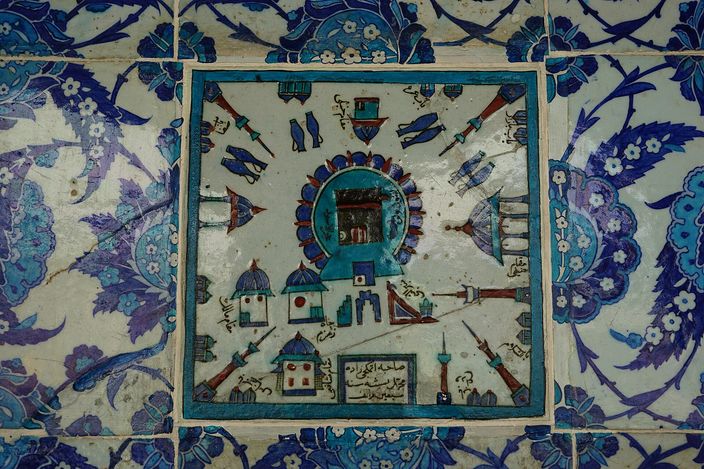 A square Iznik tile panel framed by blue-and-white floral tiles, showing a walled sanctuary in blue, red and turquoise with a dark draped cube shrine at its centre, small domed buildings and minarets around it and a cartouche of Arabic script below.
