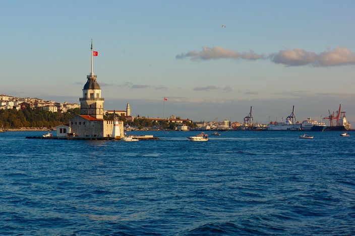A small stone tower with a conical roof and a flag standing on its own islet in a wide blue channel in evening light, motorboats crossing the water and a far shore of low buildings and port cranes behind it.
