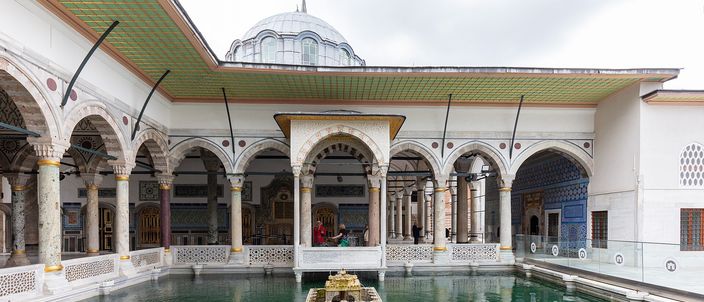 The marble terrace of an Ottoman palace: a rectangular pool of green water in front of a colonnaded arcade of pointed arches under a broad green-tiled eave, a small lead-domed pavilion rising behind it and a gilded canopy standing over the water.