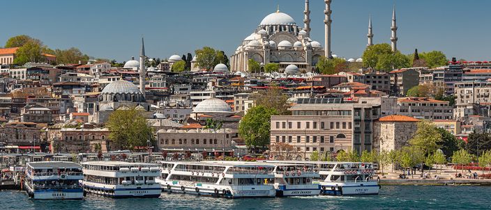 The historic peninsula from the water: the Süleymaniye Mosque and its four minarets crowning the hill above tiled roofs, with white commuter ferries moored along the Golden Horn below