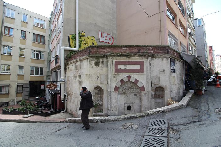 A weathered Ottoman-era stone street fountain wedged between modern apartment blocks on a sloping Cihangir side street, a man in a dark coat standing beside it.