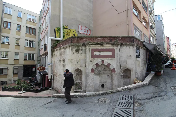 A weathered Ottoman-era stone street fountain wedged between modern apartment blocks on a sloping Cihangir side street, a man in a dark coat standing beside it.