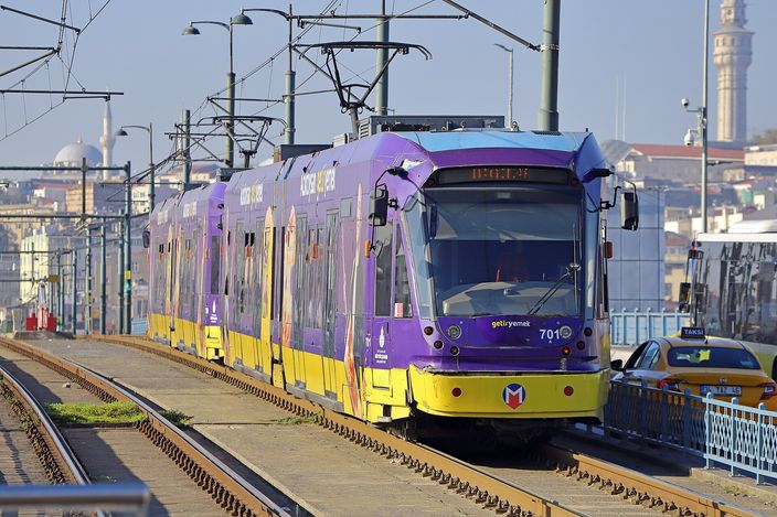 A purple-and-yellow low-floor tram of Istanbul's T1 line on its tracks by the Galata Bridge at Karaköy, with a mosque dome and minaret behind and a yellow taxi alongside