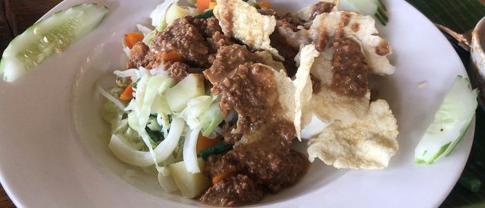 A plate of gado-gado: blanched vegetables and cucumber dressed with a thick brown peanut sauce, topped with pale crackers, on a white plate