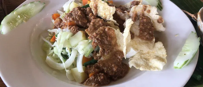 A plate of gado-gado: blanched vegetables and cucumber dressed with a thick brown peanut sauce, topped with pale crackers, on a white plate