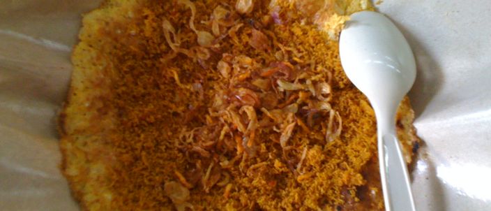 A finished kerak telor on a paper tray: a browned, crisp rice-and-egg crust topped with fried grated coconut, pounded dried shrimp and crisp fried shallots, with a spoon resting on the edge