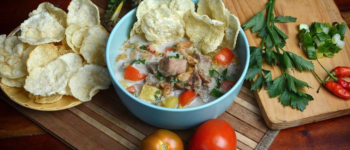 A bowl of soto Betawi, a pale creamy beef-and-offal soup, served with a wedge of tomato and a stack of melinjo crackers on a wooden board