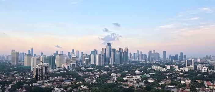 Jakarta's Sudirman/SCBD skyline of clustered high-rise towers rising above a low, green residential foreground, seen from the south at dusk under a soft pink-blue sky