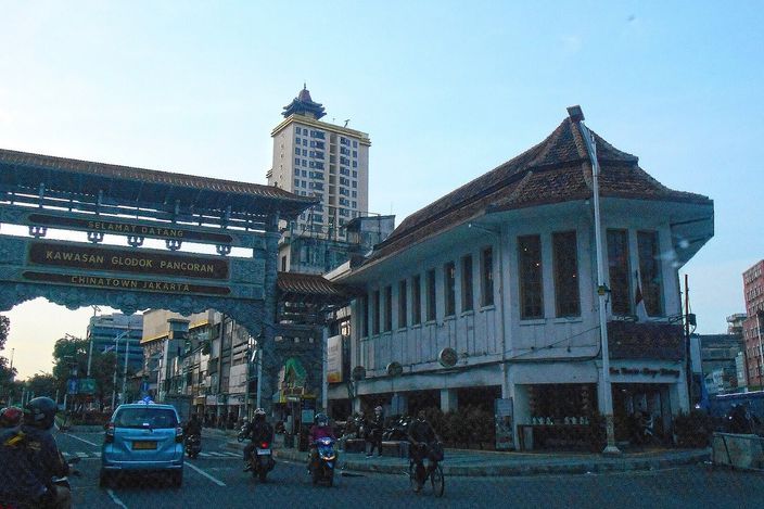 A green and gold Chinese ceremonial gateway spanning a city road, lettered "Kawasan Glodok Pancoran — Chinatown Jakarta", with an old two-storey tile-roofed shophouse beside it and motorbikes and a blue taxi passing beneath