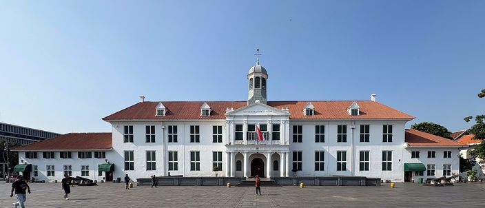 A long two-storey white colonial building with green shutters, a red tiled roof and a small clock cupola over its centre, standing behind a wide empty brick-paved square under a clear blue sky