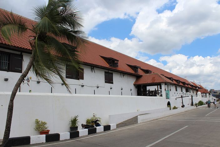 A long whitewashed Dutch East India Company warehouse range with small shuttered windows, dormers and a deep red tiled roof, running beside an empty sunlit street with a coconut palm leaning in from the left