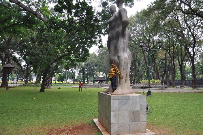 A pale stone sculpture of two entwined standing figures on a square plinth in a city park, a small child standing at its base, with big spreading rain trees, lamp posts and open lawn behind