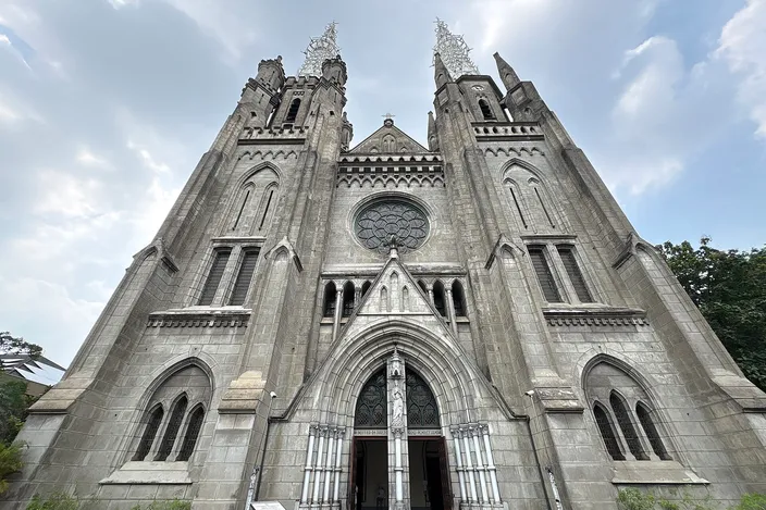 The gray stone west front of Jakarta Cathedral seen straight on: a rose window above the arched main doorway, pointed side windows, and two tall spires topped with openwork iron finials against a pale sky
