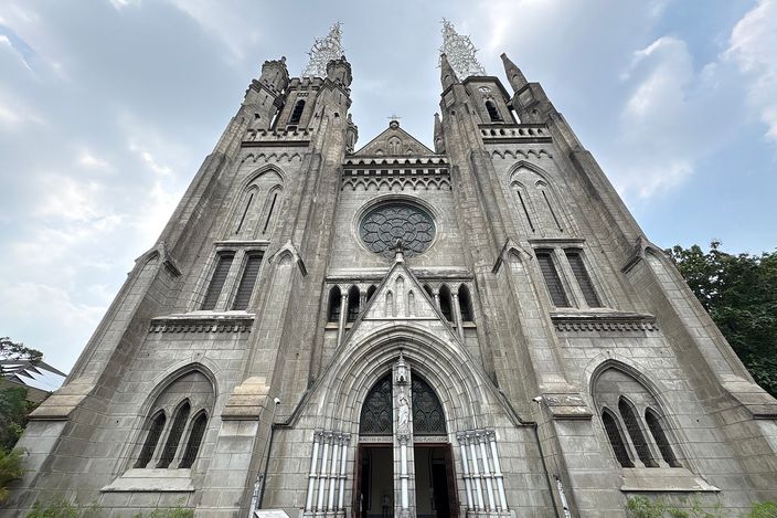 The grey stone west front of Jakarta Cathedral seen straight on: a rose window above the arched main doorway, pointed side windows, and two tall spires topped with openwork iron finials against a pale sky