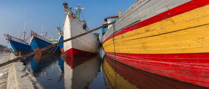Wooden pinisi cargo schooners moored bow-in along a quay at Sunda Kelapa, one hull banded red, yellow and dark red in the foreground and a line of blue-hulled ships behind, reflected in still water under a clear blue sky