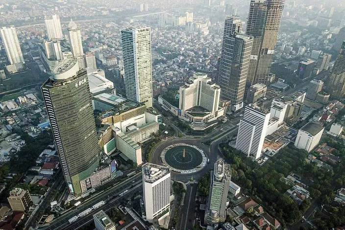 Aerial view over central Jakarta's Golden Triangle: the Hotel Indonesia roundabout with the Selamat Datang monument fountain at center, ringed by business and hotel towers fading into haze.