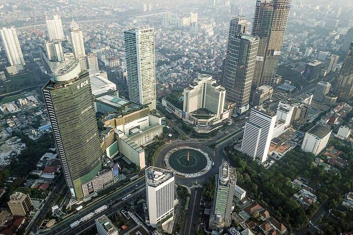 Aerial view over central Jakarta's Golden Triangle: the Hotel Indonesia roundabout with the Selamat Datang monument fountain at centre, ringed by business and hotel towers fading into haze.
