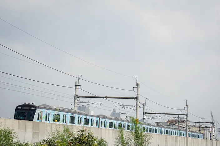 A teal-and-white MRT Jakarta train running on an elevated viaduct against a hazy sky, with overhead catenary masts and distant towers behind
